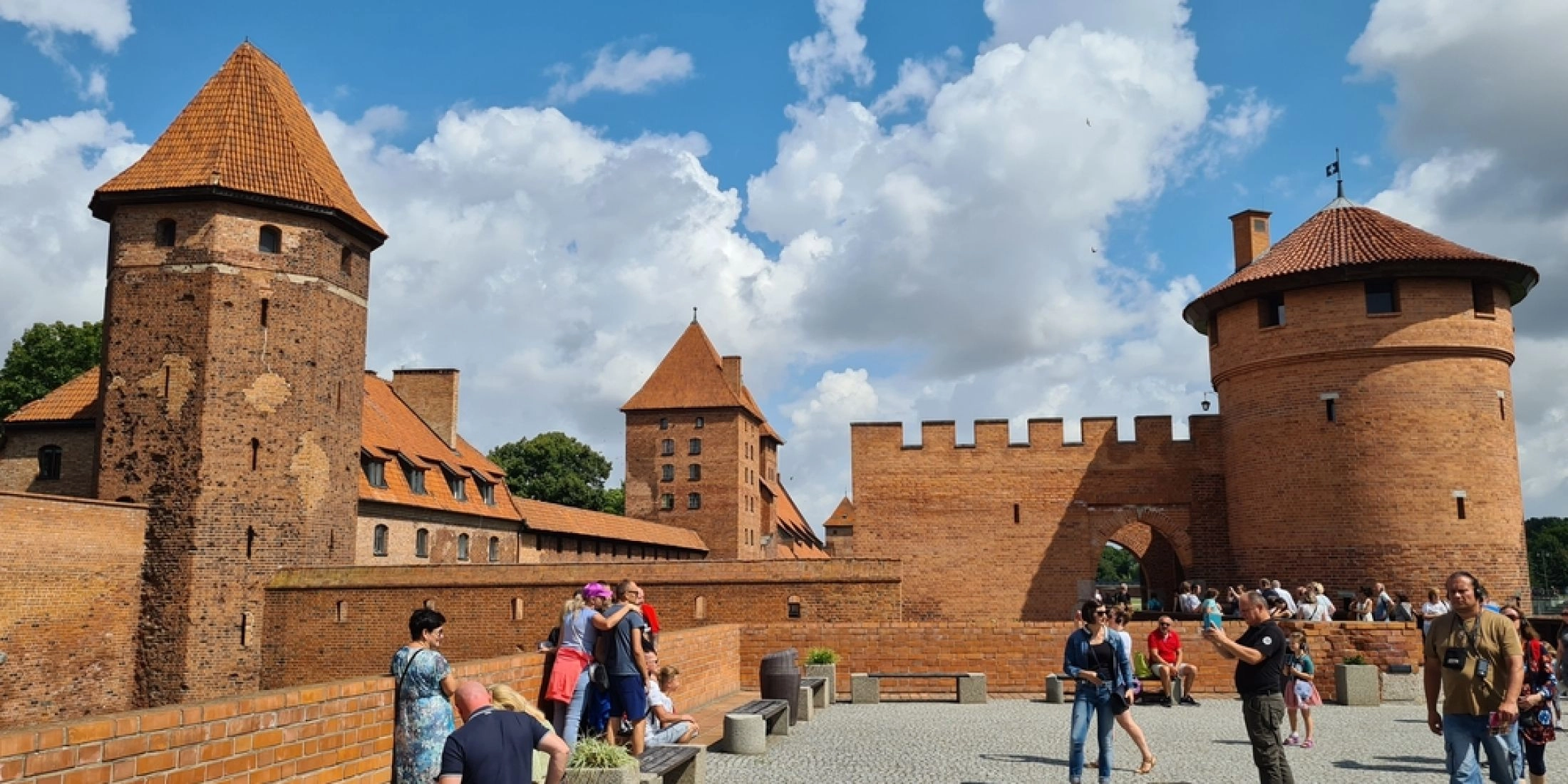 Turyści przed ceglaną bramą zamku w Malborku, otoczoną murami i basztami z dachami z czerwonej dachówki.