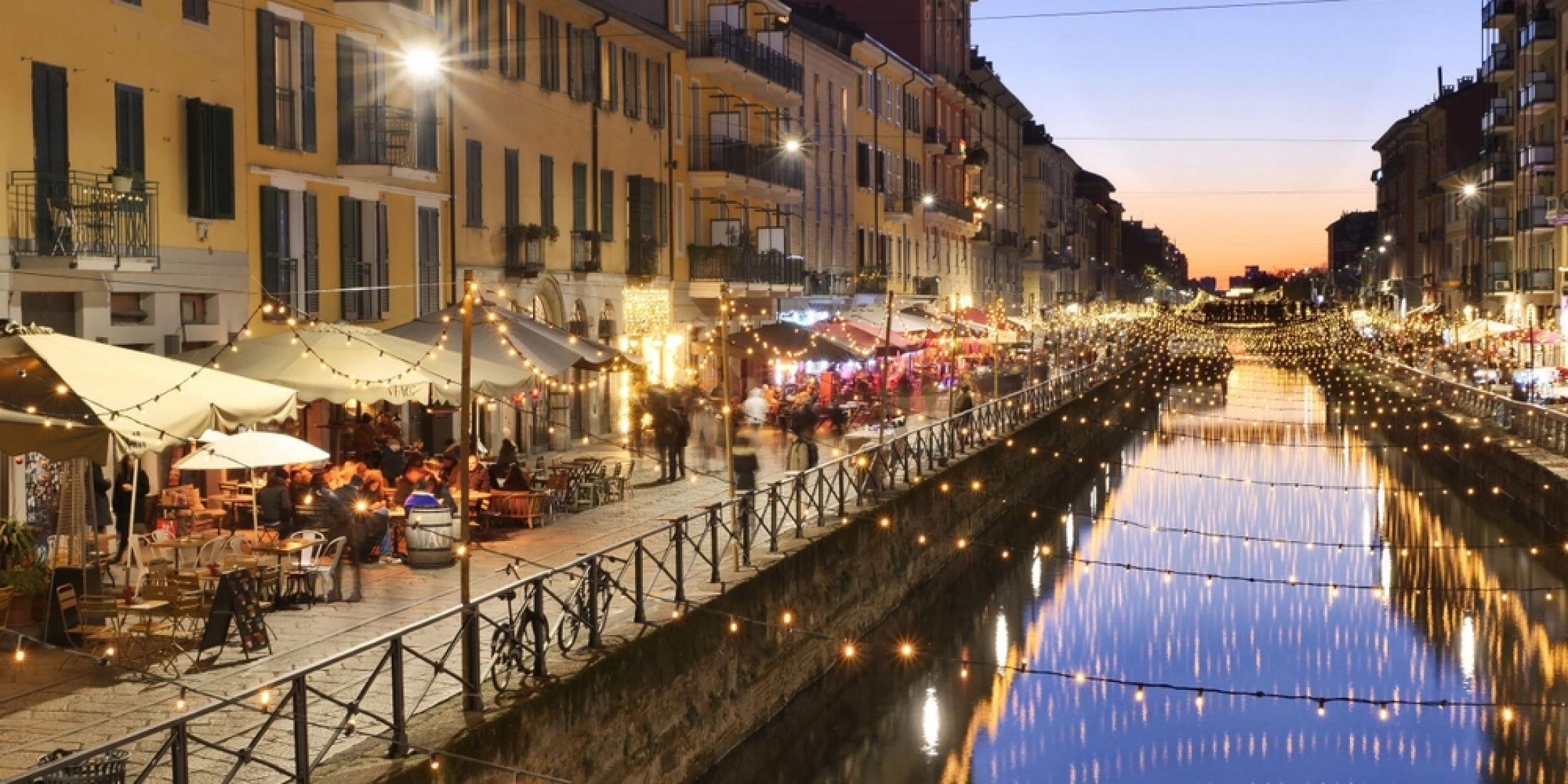 Dzielnica Navigli w Mediolanie po zmroku, z odbiciami świątecznych lampek w kanale i ogródkami restauracji.