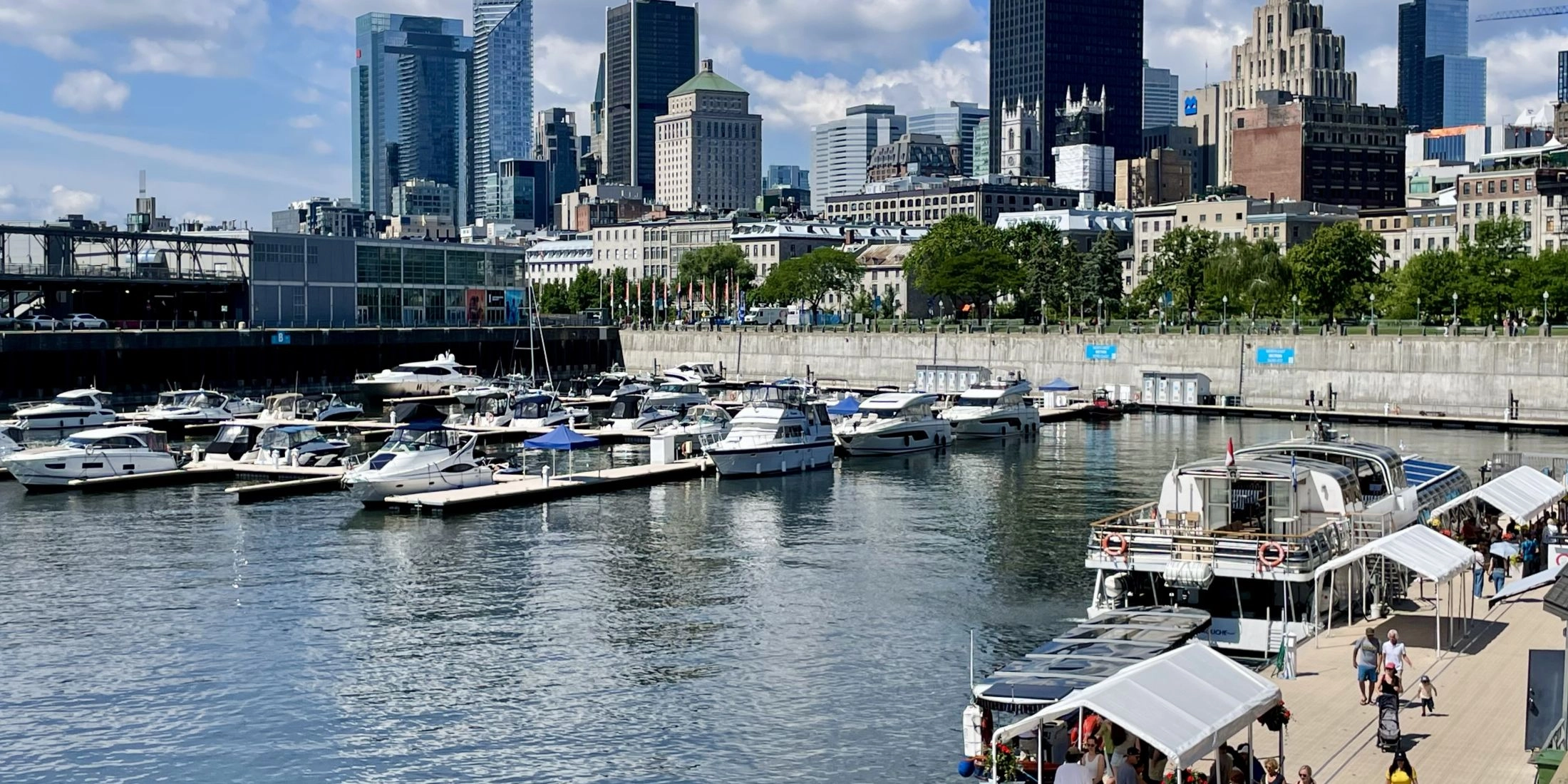 Port w Montrealu z zacumowanymi łodziami, spacerowym nabrzeżem i wieżowcami w tle.