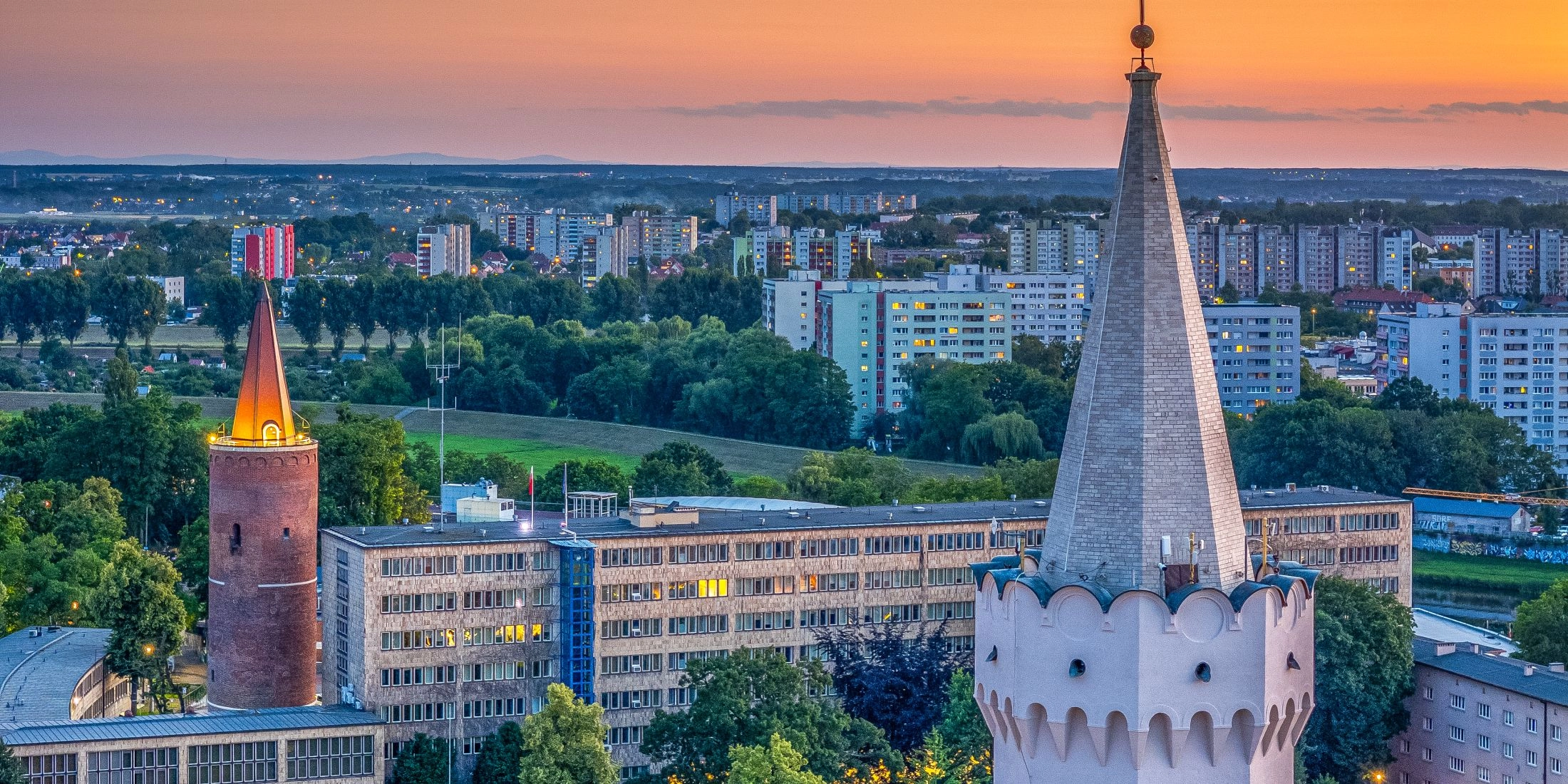 Panorama Opola o zachodzie słońca z miejskimi wieżami, zabudową centrum oraz osiedlami w tle.