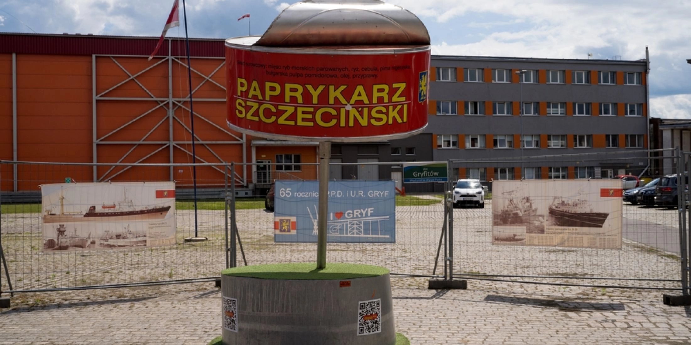 Pomnik Paprykarza Szczecińskiego w formie dużej czerwonej puszki ustawionej na okrągłym postumencie przed zabudowaniami przemysłowymi.