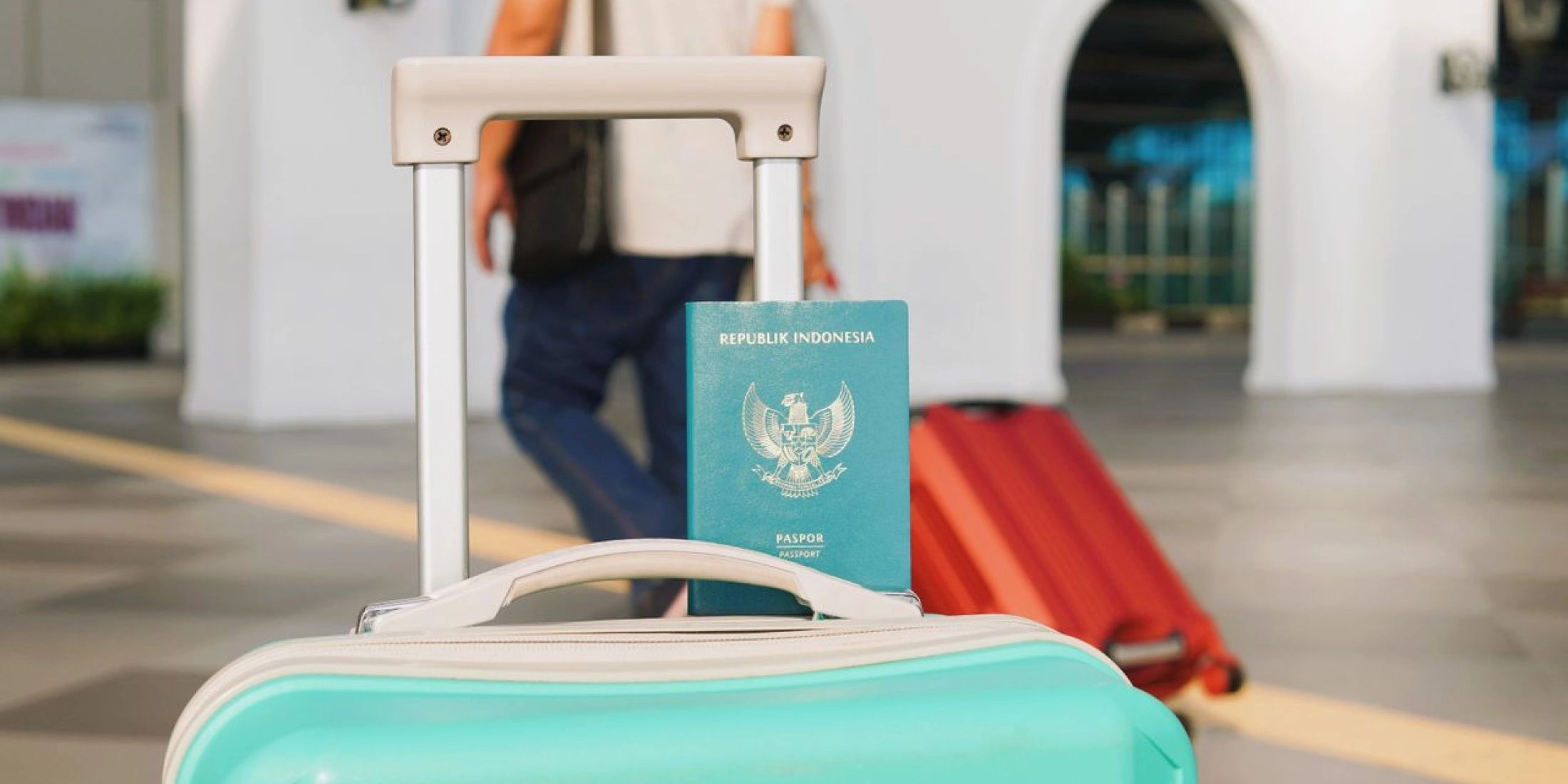 Indonezyjski paszport za uchwytem turkusowej walizki, w tle podróżny z czerwoną walizką.