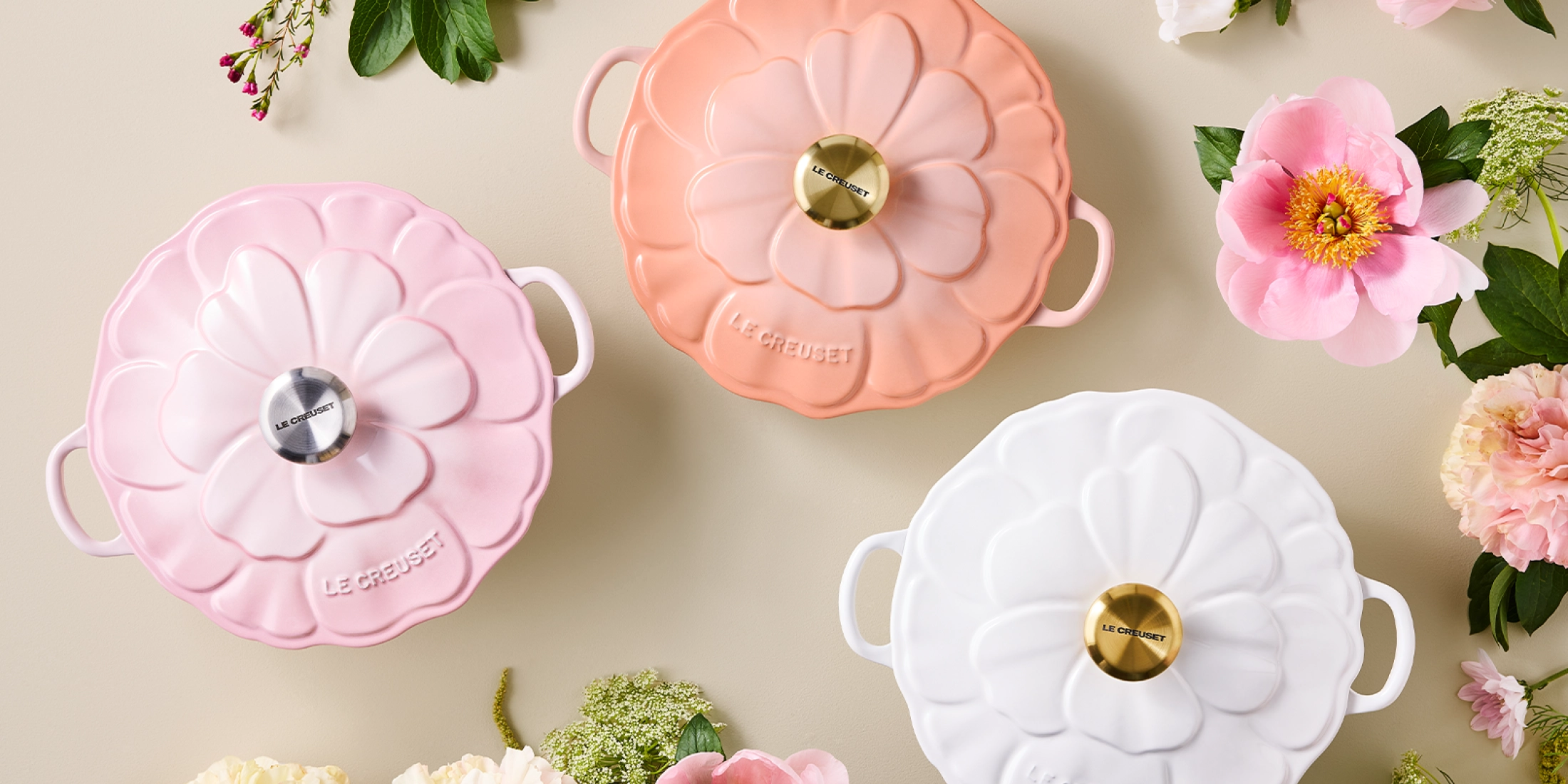Trzy pastelowe naczynia Le Creuset w kształcie kwiatu, otoczone świeżymi kwiatami na jasnym tle.