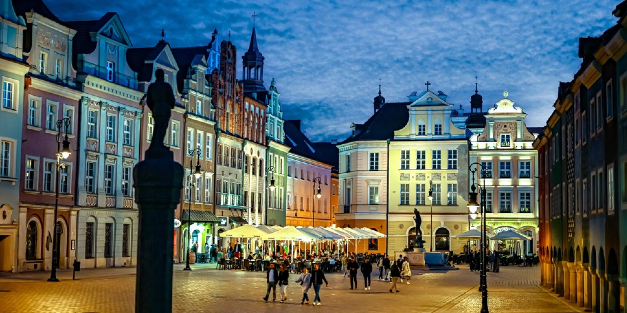 Stary Rynek w Poznaniu nocą z podświetlonymi kamienicami i spacerującymi ludźmi.