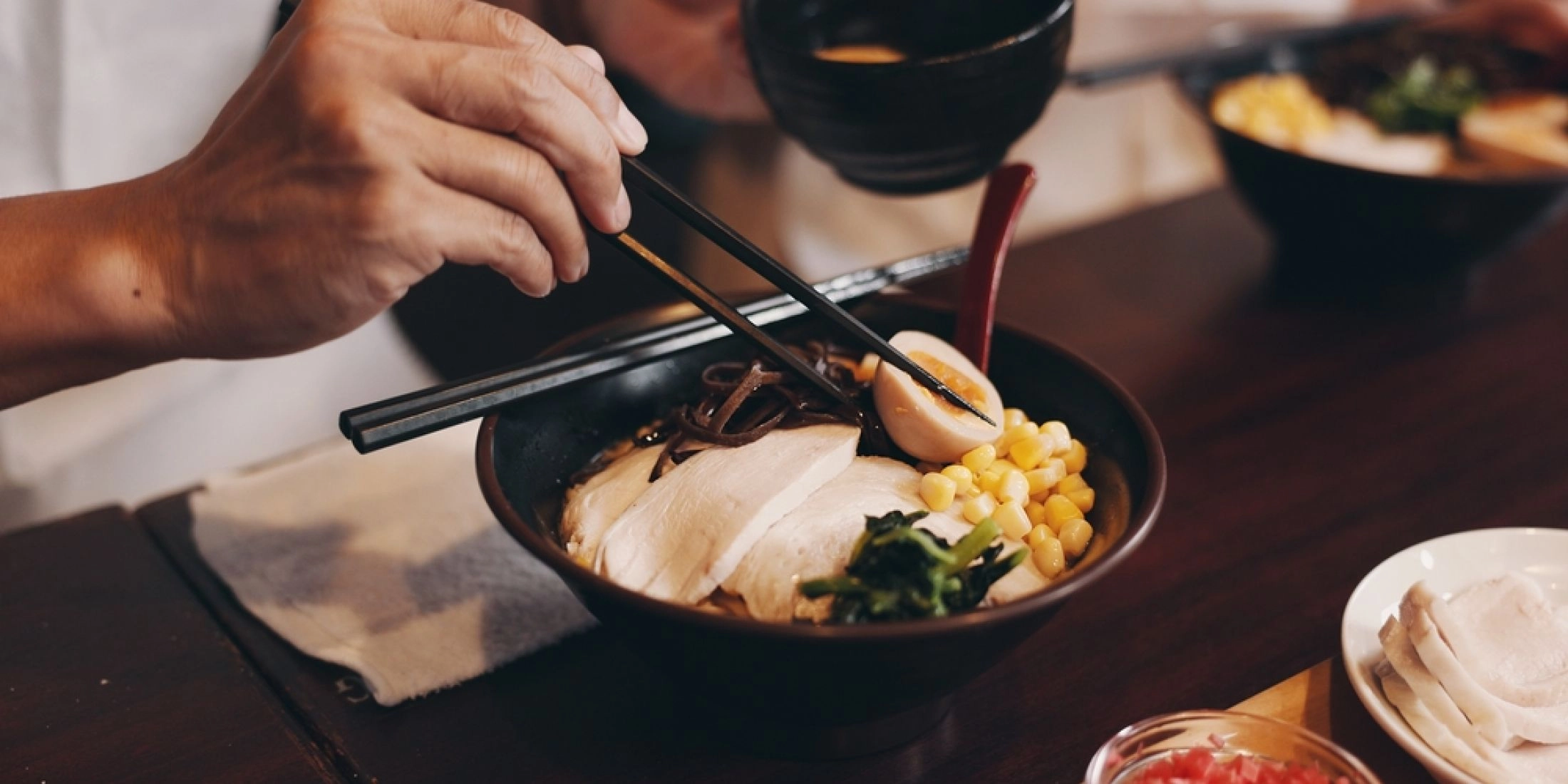 Osoba je ramen pałeczkami z czarnej miski, w której widać plastry mięsa, jajko, kukurydzę i warzywa.