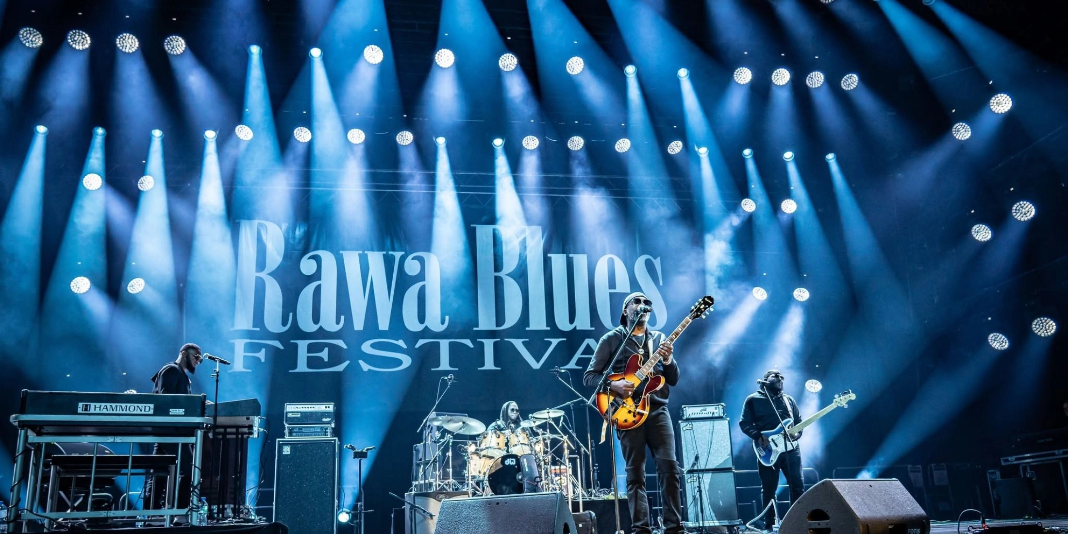 Gitarzysta przy mikrofonie, klawiszowiec, perkusista i basista na scenie Rawa Blues Festival oświetlonej niebieskimi reflektorami.
