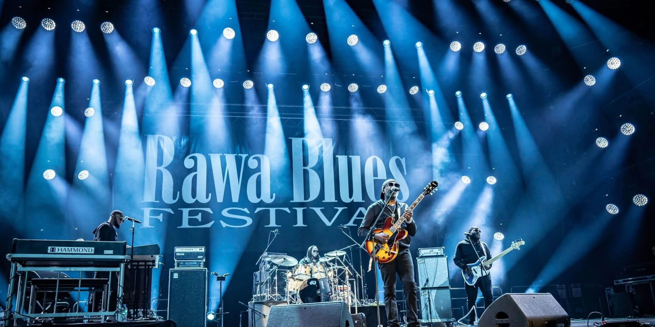 Gitarzysta przy mikrofonie, klawiszowiec, perkusista i basista na scenie Rawa Blues Festival oświetlonej niebieskimi reflektorami.