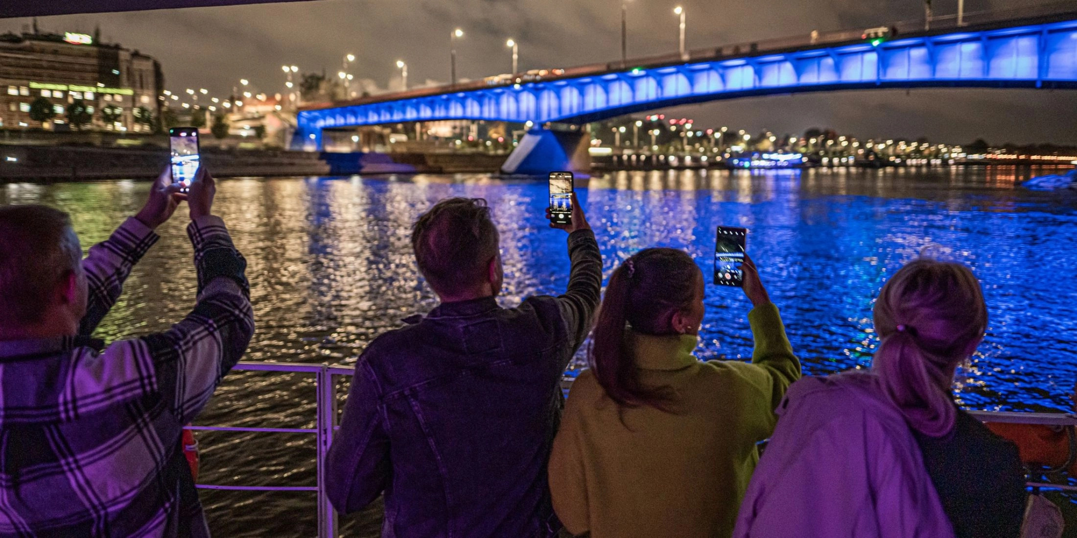 Uczestnicy rejsu fotografują nocną Wisłę i podświetlony most podczas wydarzenia Riverlights w Warszawie.