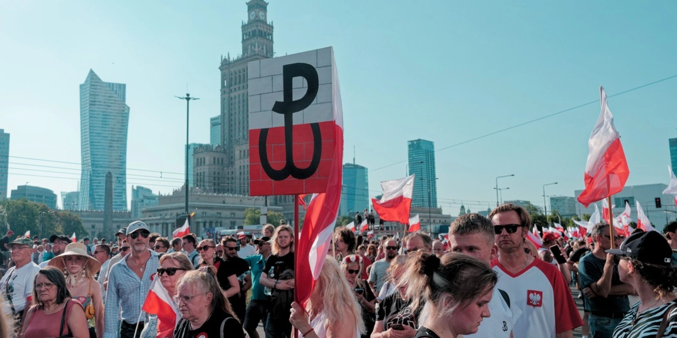 Tłum uczestników obchodów Powstania Warszawskiego z biało-czerwonymi flagami i znakiem Polski Walczącej w centrum Warszawy.