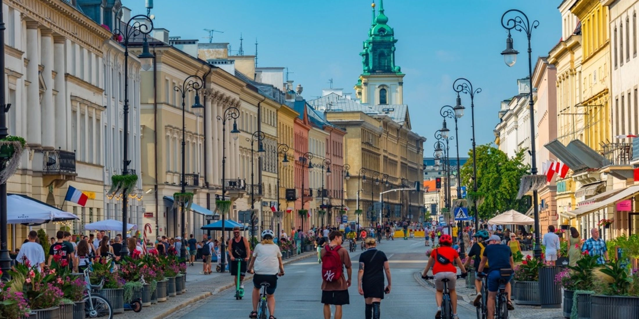 Spacerowicze i rowerzyści na Krakowskim Przedmieściu w Warszawie, w tle kolorowe kamienice i wieża kościoła.