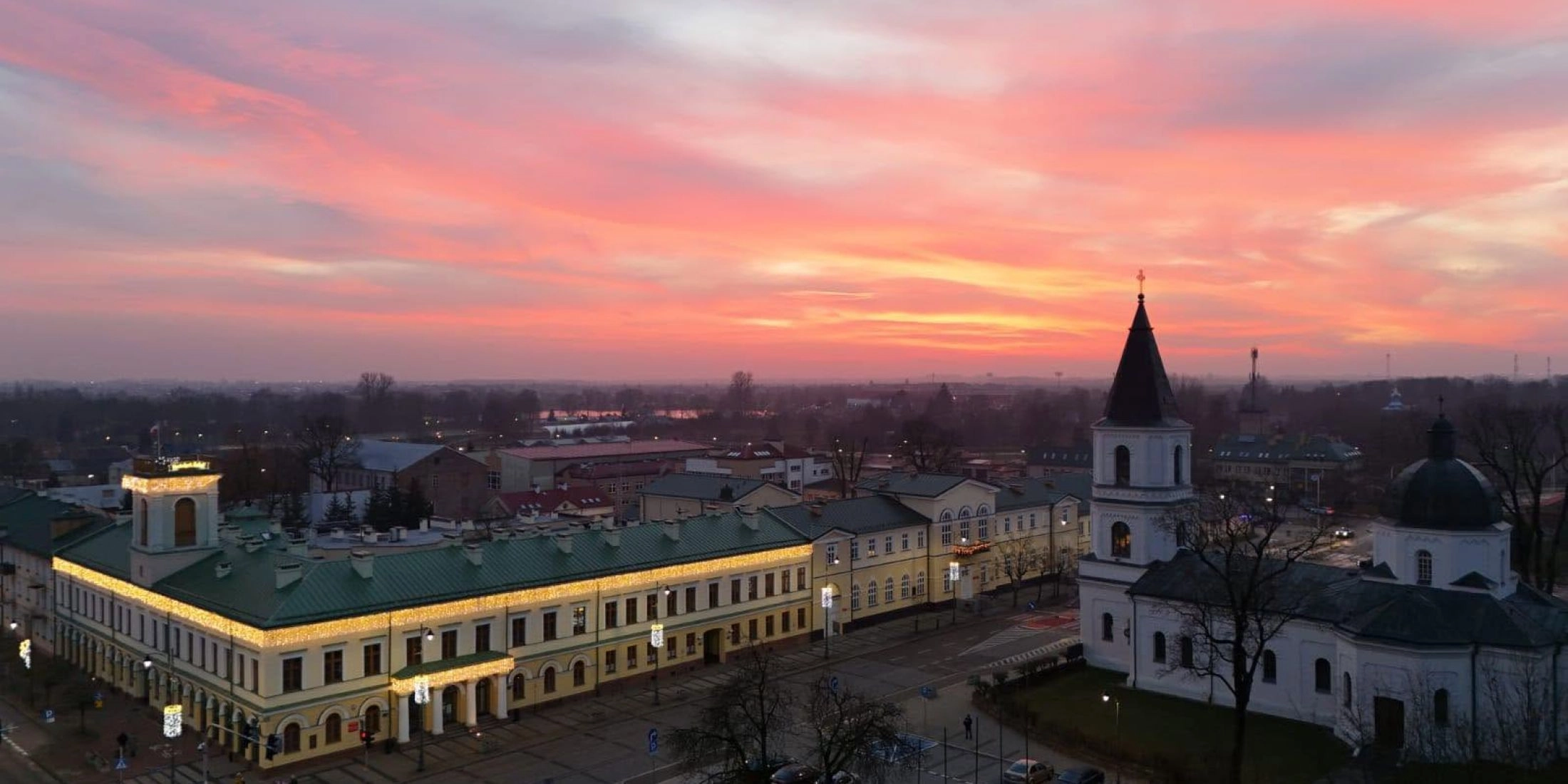 Centrum Suwałk widziane z góry o zachodzie słońca, z zabytkową zabudową, kościołem i różowym niebem nad miastem.