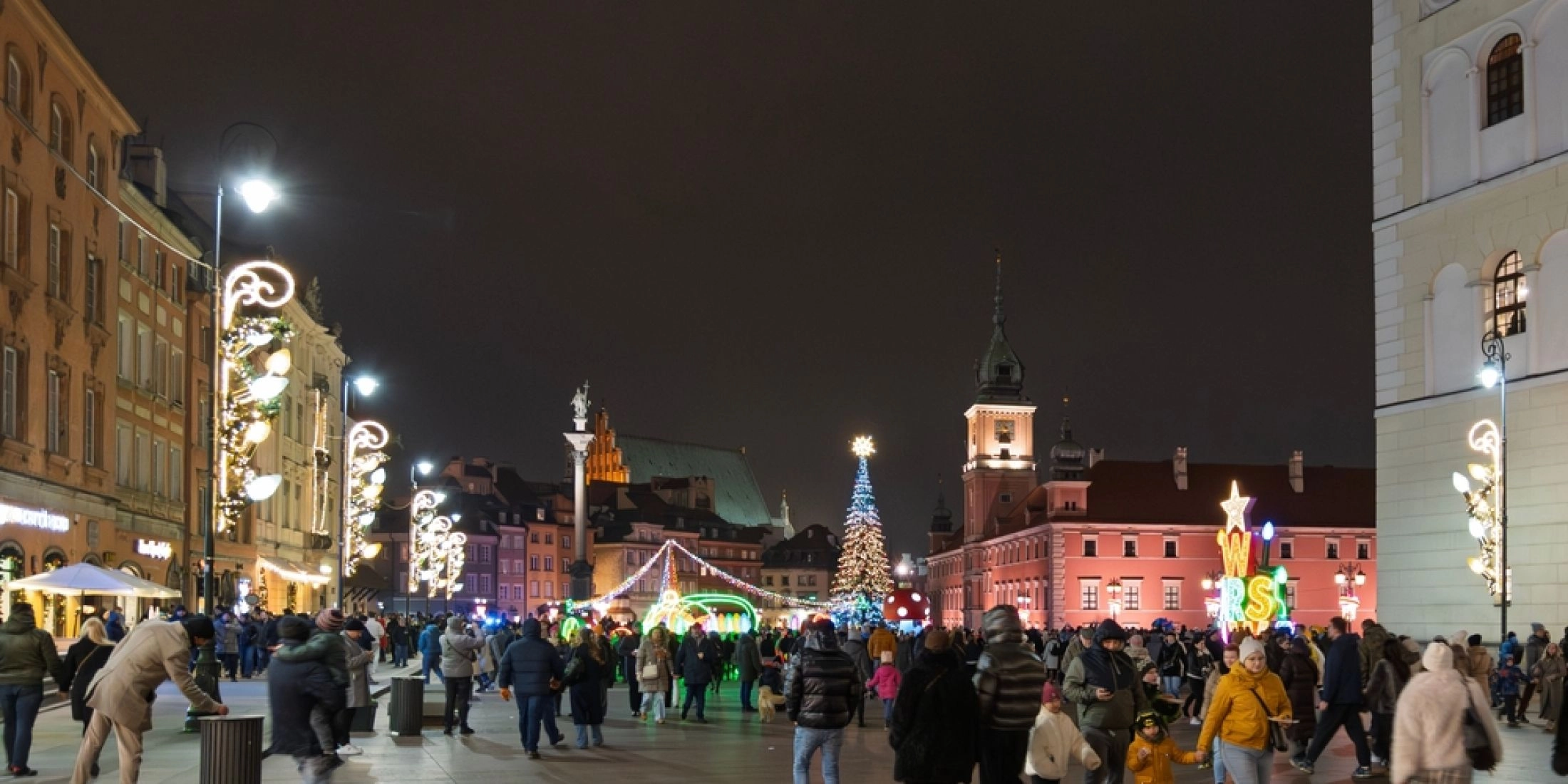 Świąteczny Plac Zamkowy w Warszawie nocą: choinka, iluminacje i spacerujący ludzie na starówce zimą.