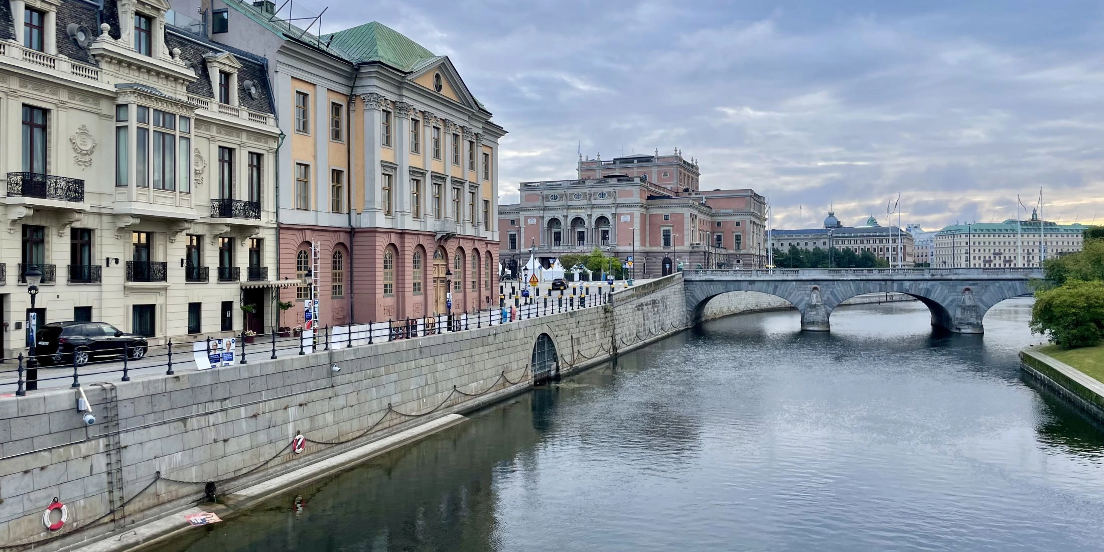Zabytkowe budynki nad wodą w centrum Sztokholmu, z kamiennym mostem i gmachem Opery Królewskiej w tle.