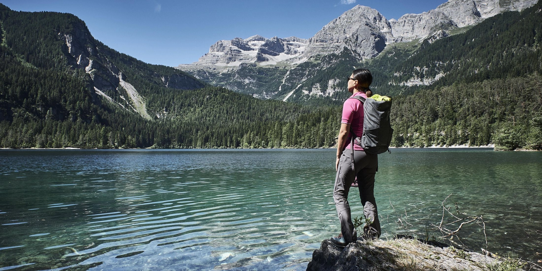 Turystka z plecakiem stoi nad górskim jeziorem w Trentino, w tle widać lasy i skaliste szczyty Dolomitów.