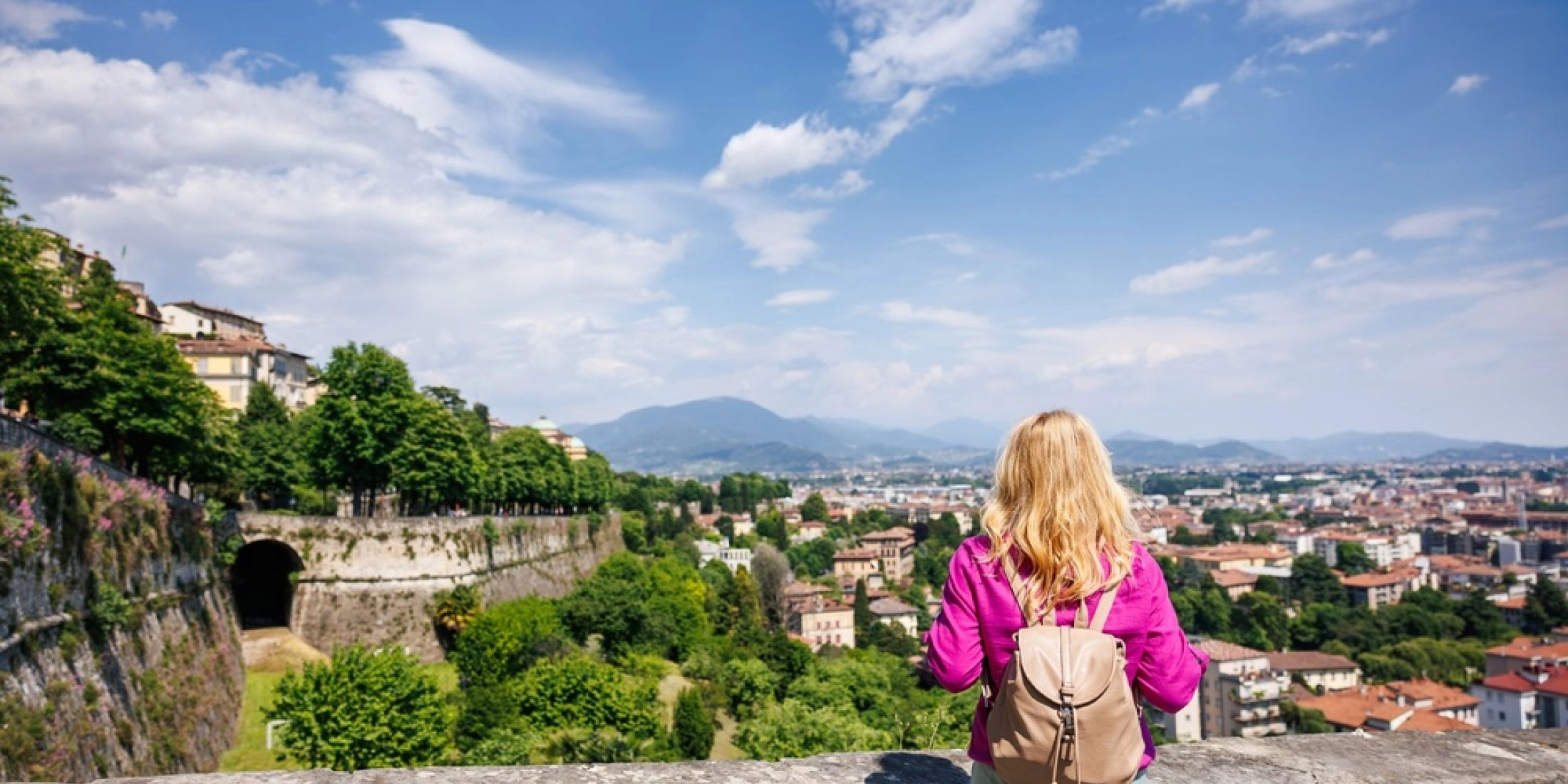 Turystka z plecakiem stojąca przy kamiennym murze i oglądająca panoramę Bergamo oraz okoliczne wzgórza.