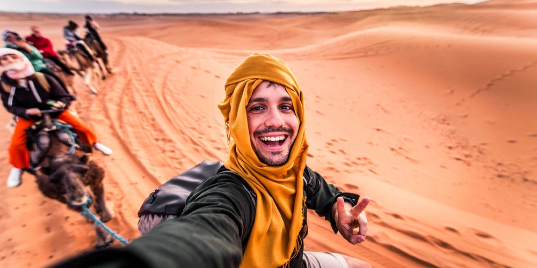 Mężczyzna w żółtym chustowym zawoju robiący selfie podczas wycieczki na wielbłądach po piaszczystej pustyni w Maroku.