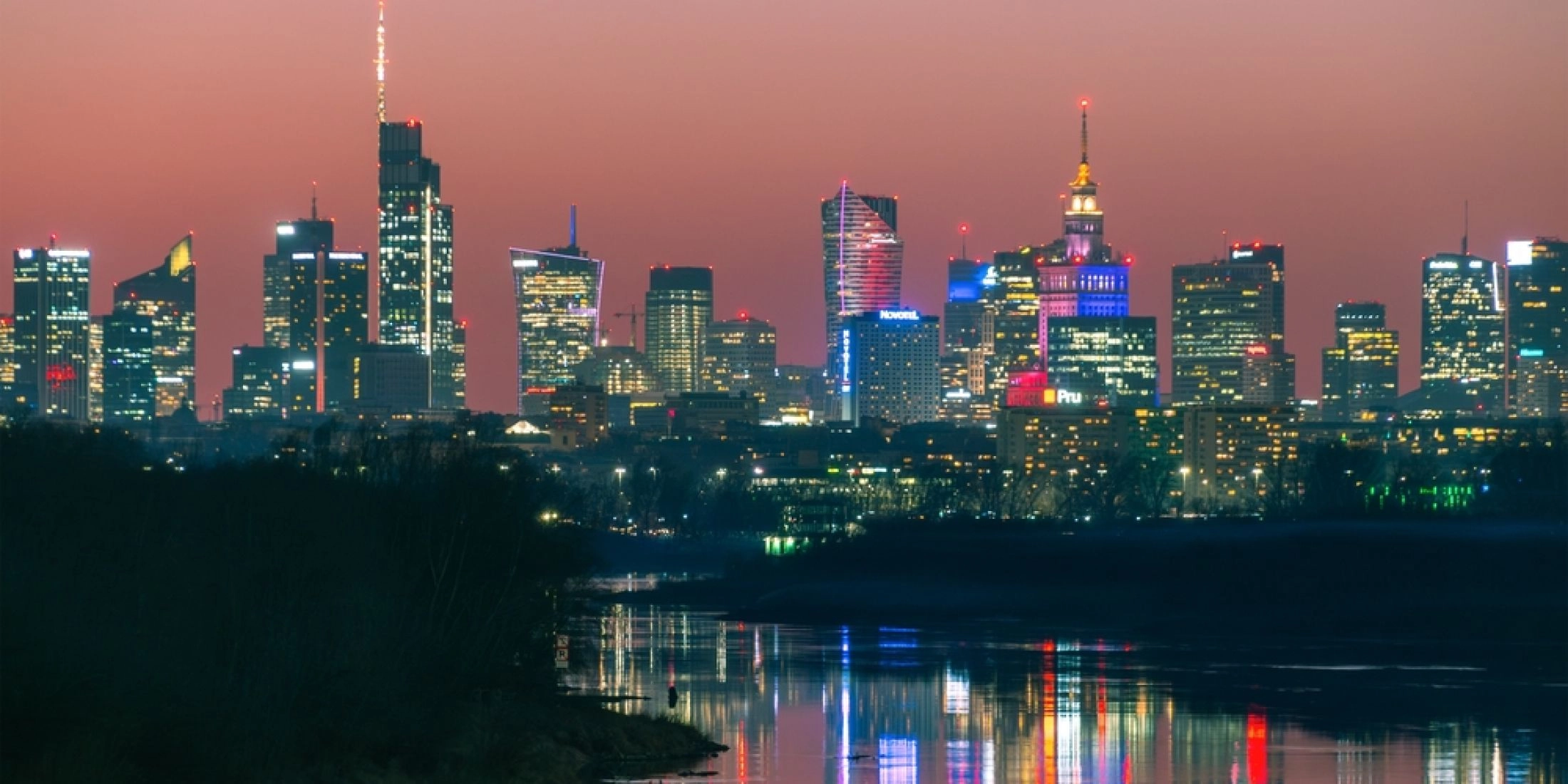 Panorama centrum Warszawy nocą z widokiem na Pałac Kultury i Nauki i wieżowce odbijające się w Wiśle.