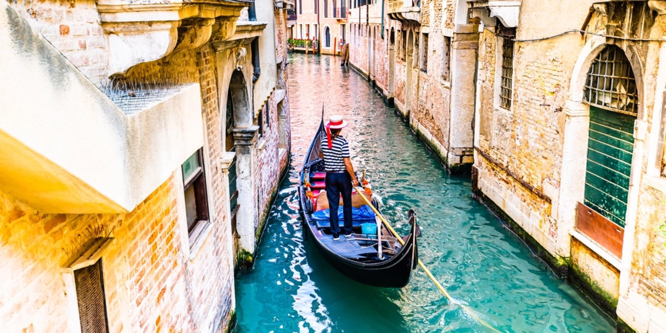 Gondola w wąskim kanale w Wenecji, gondolier w czerwonym kapeluszu między zabytkowymi kamienicami
