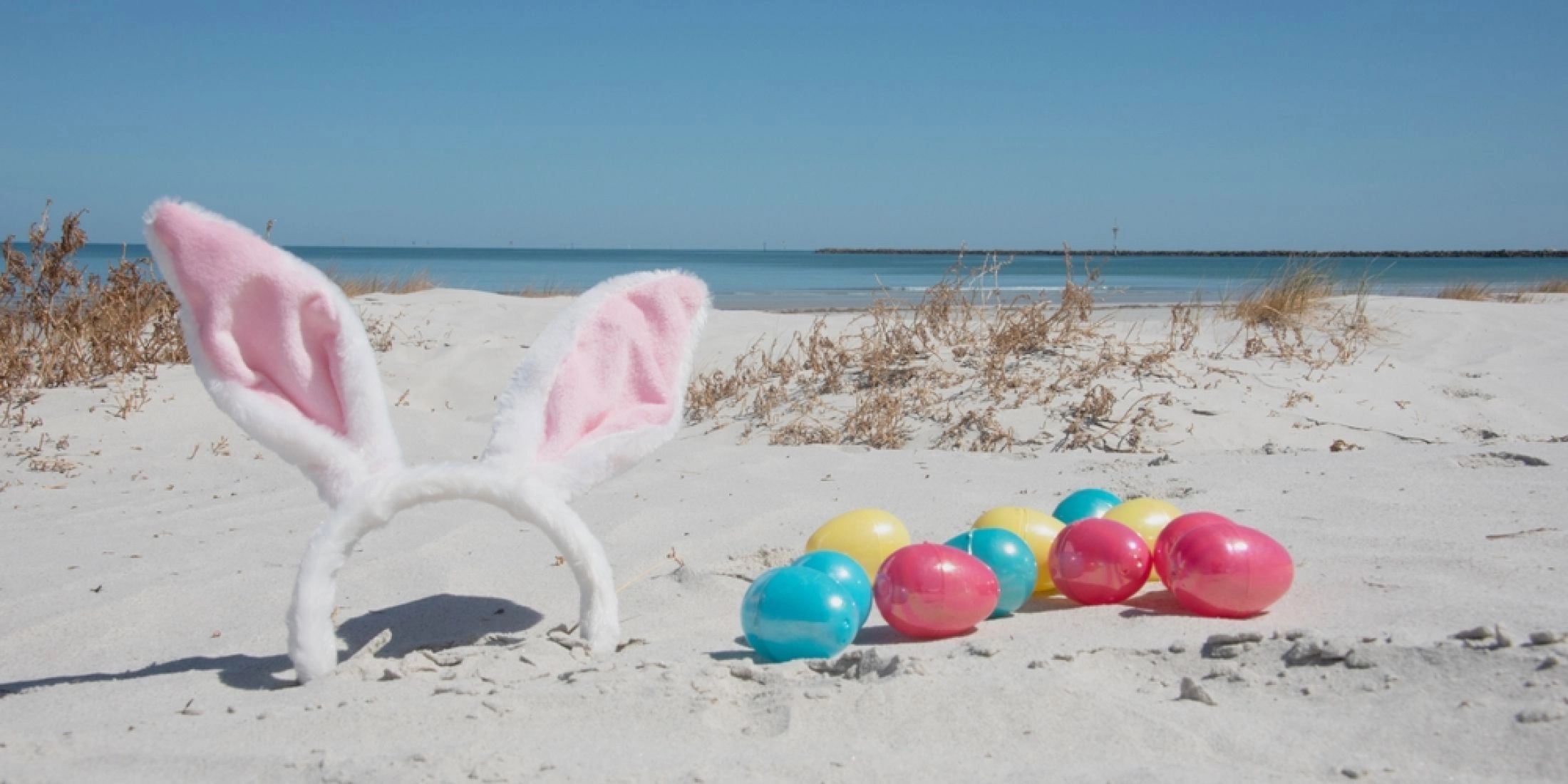 Kolorowe wielkanocne jajka i opaska z uszami królika ustawione na jasnym piasku przy plaży nad morzem.