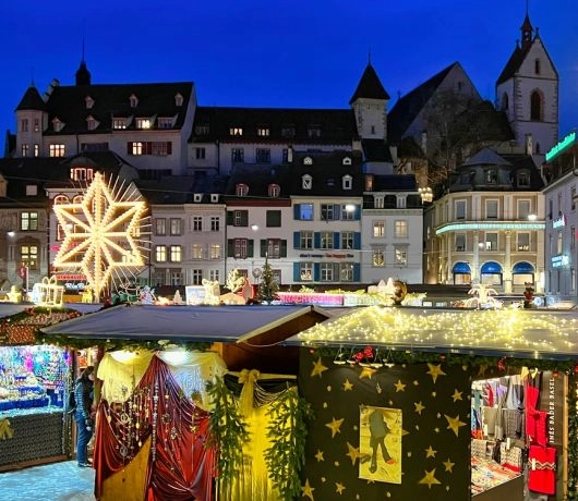 Wieczorny jarmark w Bazylei z oświetlonymi stoiskami i kamienicami, dekoracje gwiazd i świąteczne światła.