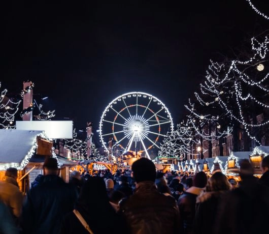 Nocny kadr z jarmarku w Brukseli z diabelskim młynem w tle i aleją straganów pełnych odwiedzających.