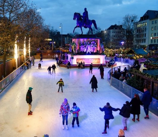 Lodowisko przy jarmarku w Kolonii z pomnikiem jeźdźca i świątecznymi dekoracjami, ludzie jeżdżą na łyżwach.