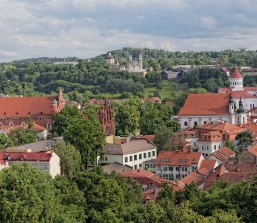 Współczesna panorama Wilna z Góry Zamkowej z czerwonymi dachami, zabytkowymi kościołami i drzewami otaczającymi zabudowę.