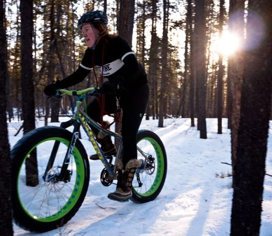 Kobieta jedzie na rowerze typu fatbike z szerokimi oponami przez ośnieżony sosnowy las.
