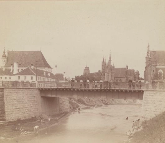 Most na Wilejce w Wilnie na archiwalnej fotografii, z ludźmi na moście i nad wodą oraz kościołami i Górą Zamkową w tle.