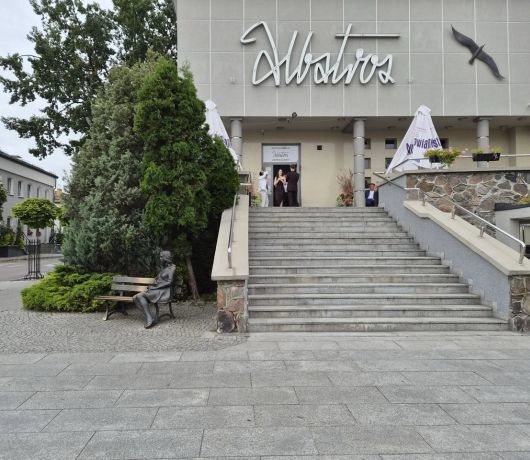 Wejście do restauracji Albatros w Augustowie, szerokie schody i pomnik Beaty siedzącej na ławce obok budynku.