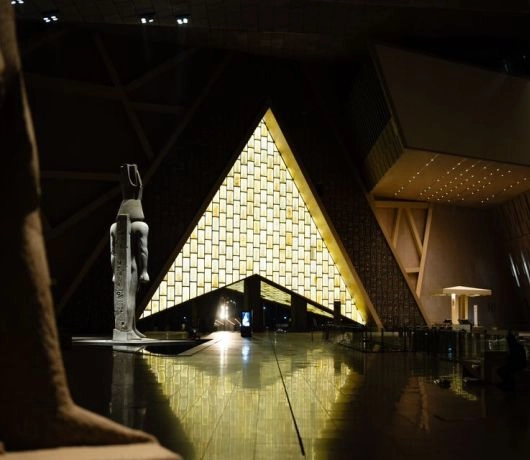 Oświetlone wejście Grand Egyptian Museum z geometryczną konstrukcją przypominającą piramidę.