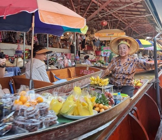Kobieta na pływającym targu w Bangkoku sprzedająca owoce z łodzi, otoczona turystami pod kolorowymi parasolami.