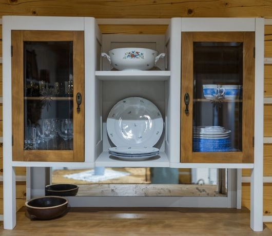 Biały drewniany kredens ze szklanymi drzwiczkami, starymi naczyniami i porcelaną na tle ściany z drewnianych bali.
