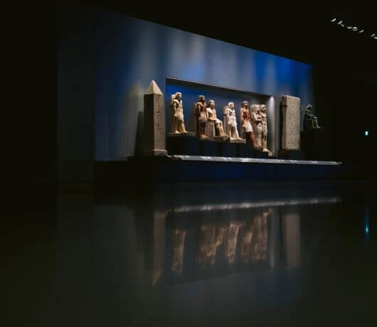 Długa sala wystawowa z figurami i obeliskiem odbijającymi się w połyskującej posadzce.