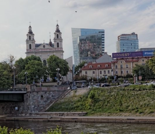 Widok przez Wilię na kościół św. Rafała, drzewa i nowoczesne biurowce, z mostem po lewej stronie.