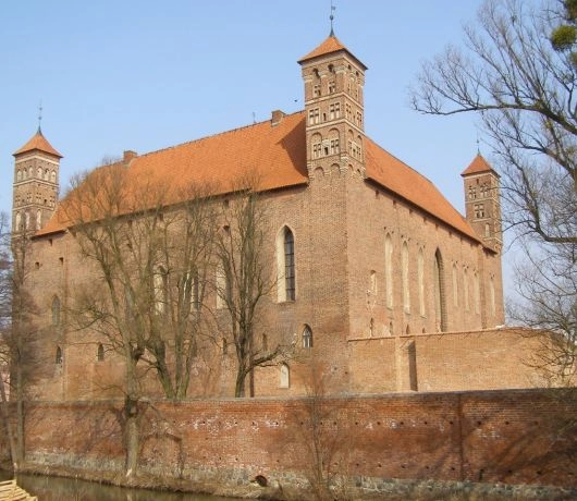 otycki Zamek Biskupów Warmińskich w Lidzbarku Warmińskim z ceglanymi murami, wieżami i charakterystycznym czerwonym dachem.