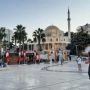 Plac w albańskim mieście z meczetem, palmami, ogródkami kawiarni i spacerującymi ludźmi o zmierzchu.