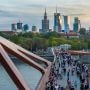 Tłum spacerowiczów i rowerzystów na kładce pieszo-rowerowej nad Wisłą w Warszawie, z panoramą centrum w tle.