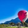 Różowy balon w kształcie głowy kota lecący nisko nad górską doliną, z ośnieżonymi szczytami w tle.
