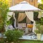 Pergola ogrodowa z jasnym dachem, zasłonami, poduszkami i dekoracjami ustawiona na drewnianym podestie wśród roślin.