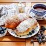 Dwie jagodzianki posypane cukrem pudrem na talerzu, podane z filiżanką herbaty i świeżymi jagodami.
