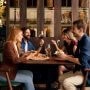 Cztery osoby siedzą przy stole w restauracji, jedzą i rozmawiają w przytulnym wnętrzu z drewnianą zabudową i półkami z przetworami w tle.