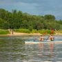 Dwie osoby w kamizelkach ratunkowych płyną dwuosobowym kajakiem po Wiśle, w tle piaszczysta plaża i zielony brzeg.