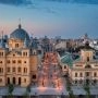 Widok z lotu ptaka na Plac Wolności i początek ulicy Piotrkowskiej w Łodzi o zmierzchu. Po lewej stronie widać charakterystyczny budynek kościoła Zesłania Ducha Świętego z kopułą, a po prawej budynek z wieżyczką zegarową. Ulica Piotrkowska jest oświetlona