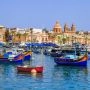 Kolorowe łodzie luzzu w porcie Marsaxlokk na Malcie, w tle zabudowa miasteczka i kościół.