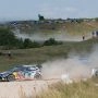 Samochód rajdowy Volkswagen pokonuje szutrowy zakręt na pagórkowatej trasie obserwowanej przez licznych kibiców.