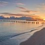 Spokojna plaża nad Bałtykiem o zachodzie słońca, z drewnianymi palami w wodzie i łagodną linią brzegu.