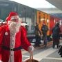 Mikołaj z koszem prezentów na peronie obok świątecznego pociągu w kolorowych wagonach, w tle pasażerowie w zimowych ubraniach.
