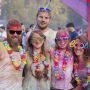Grupa pięciu uśmiechniętych uczestników festiwalu w kolorowych okularach, kwiatowych girlandach i ubraniach pokrytych barwnym proszkiem.