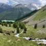 Górski szlak trekkingowy w Val Senales z potokiem, zielonymi halami i ośnieżonymi szczytami w tle.